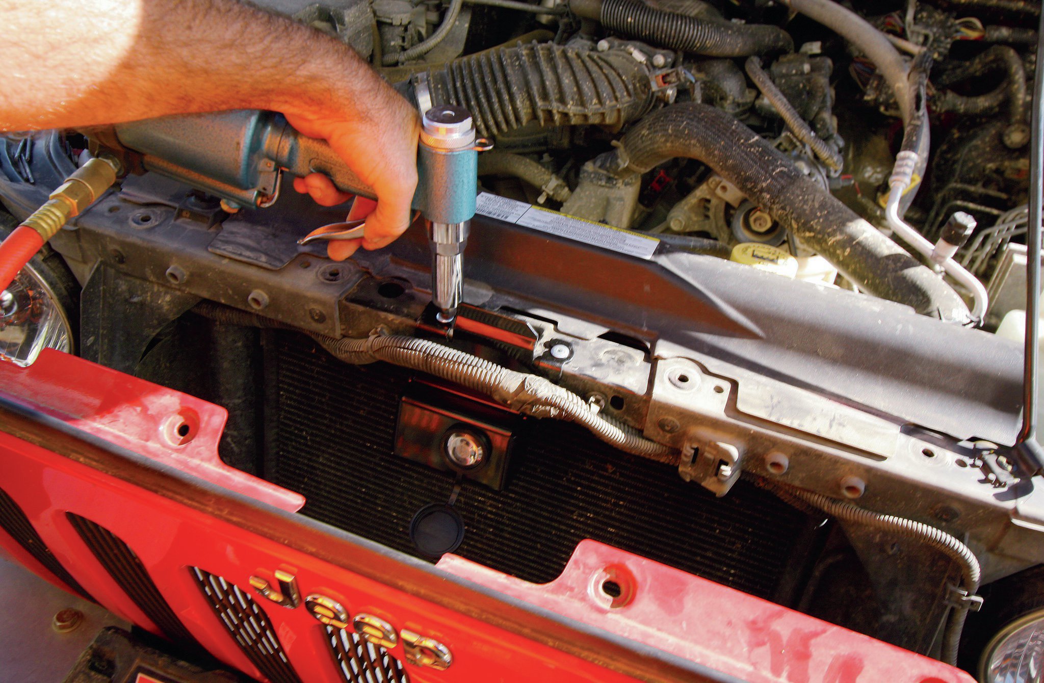 Use a small flat-head screwdriver to pry up the plastic clips holding the grill on. Don’t worry too much about breaking the clips—new ones are included with the hood lock. We found that it wasn’t necessary to remove the entire grill. Letting it lean forward against our winch control box worked just fine to keep it out of the way.